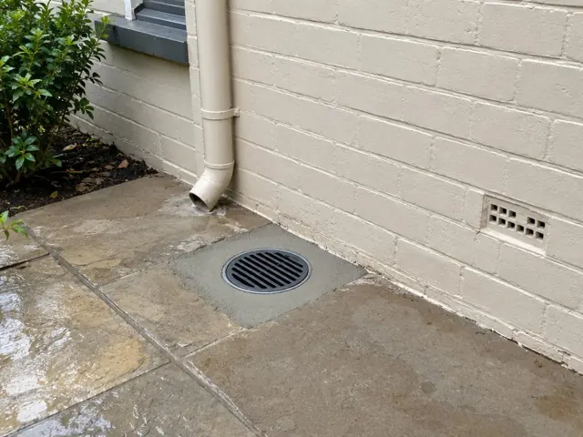 Clean outdoor drain grate beside a cream brick wall and downpipe on wet paving in Bellevue Hill